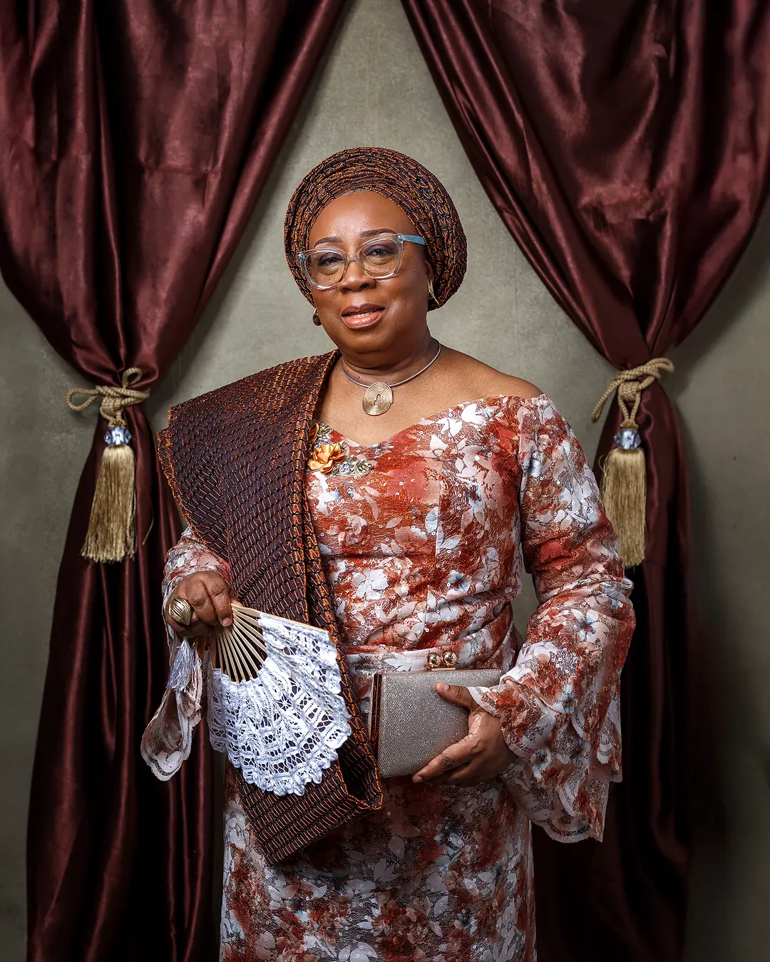 Folasade Adefisayo in a formal patterned gown and gele, holding a fan, between burgundy drapes.