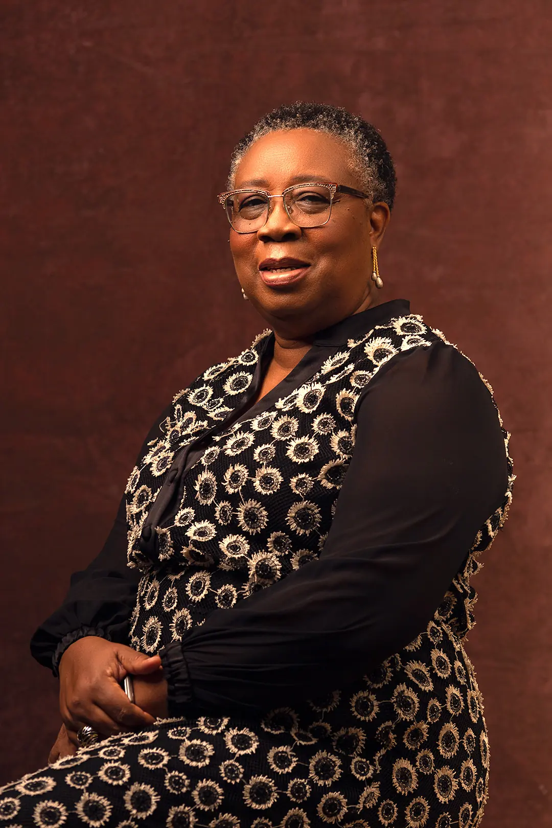 Folasade Adefisayo seated in a patterned black and gold outfit, arm resting on a stool.