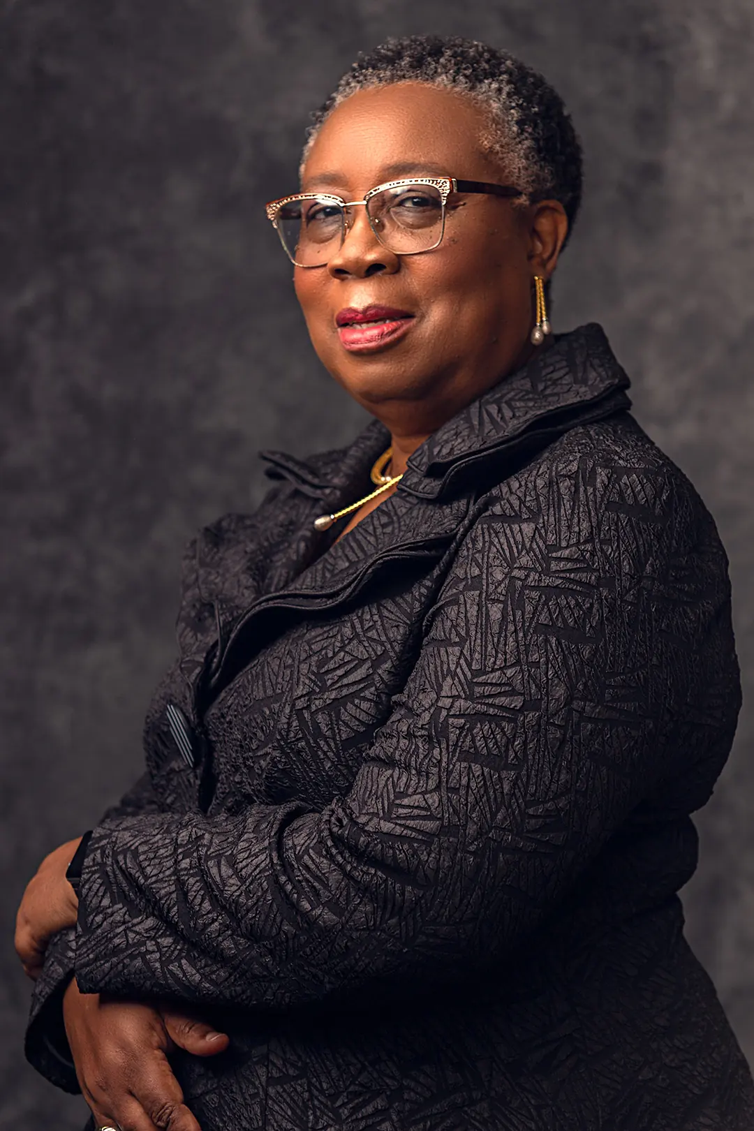 Folasade Adefisayo standing with arms folded in a dark jacket against a grey backdrop.